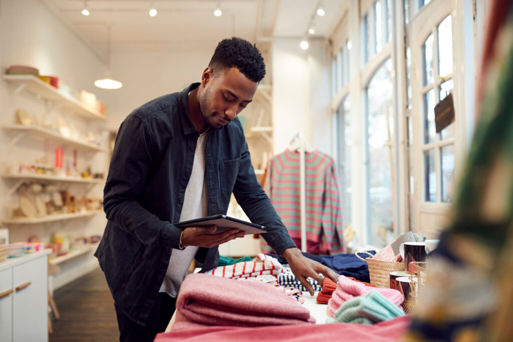 A Black male associate reviews a tablet over a merchandising table in a boutique — store execution, tasks, merchandising.