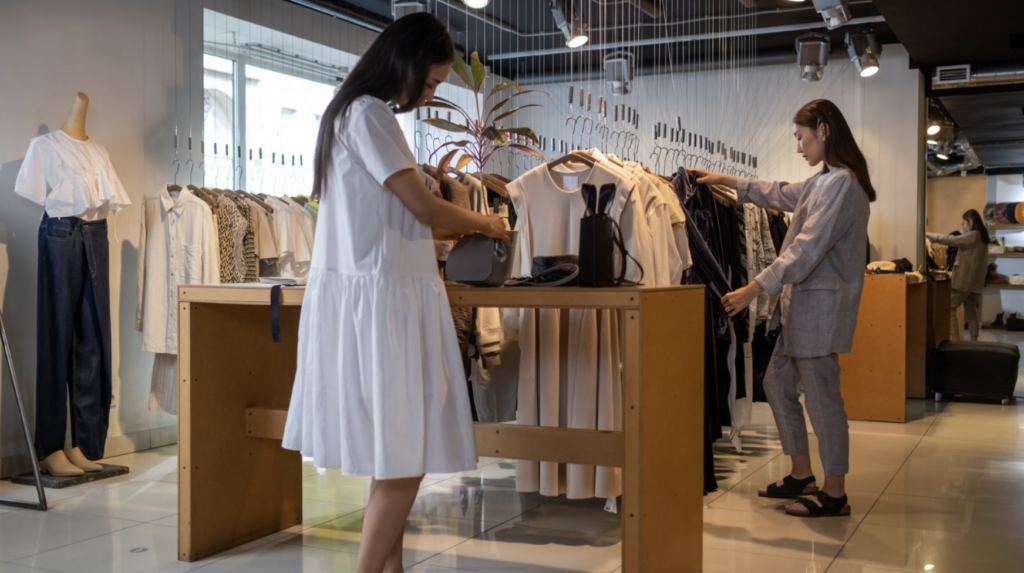 Two shoppers looking through neutral-toned clothing on racks and a central display table in a bright, minimalist store.