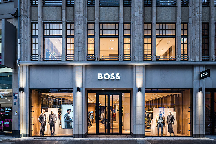 Exterior of a BOSS store lit up in the evening, with menswear and womenswear displayed across large window fronts.
