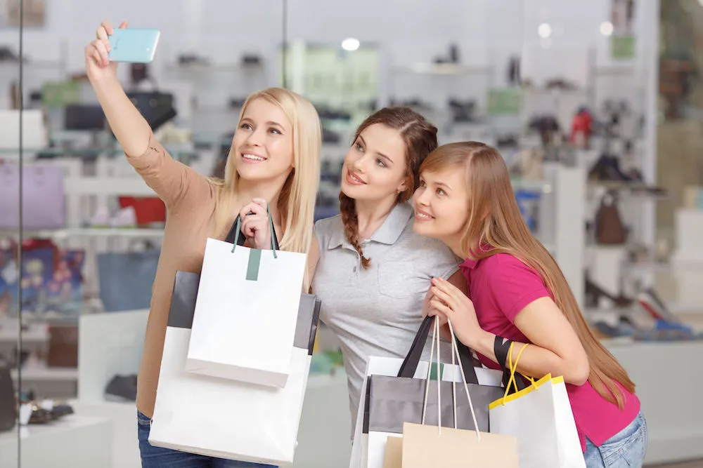 Three young shoppers taking a selfie together inside a store while holding shopping bags.