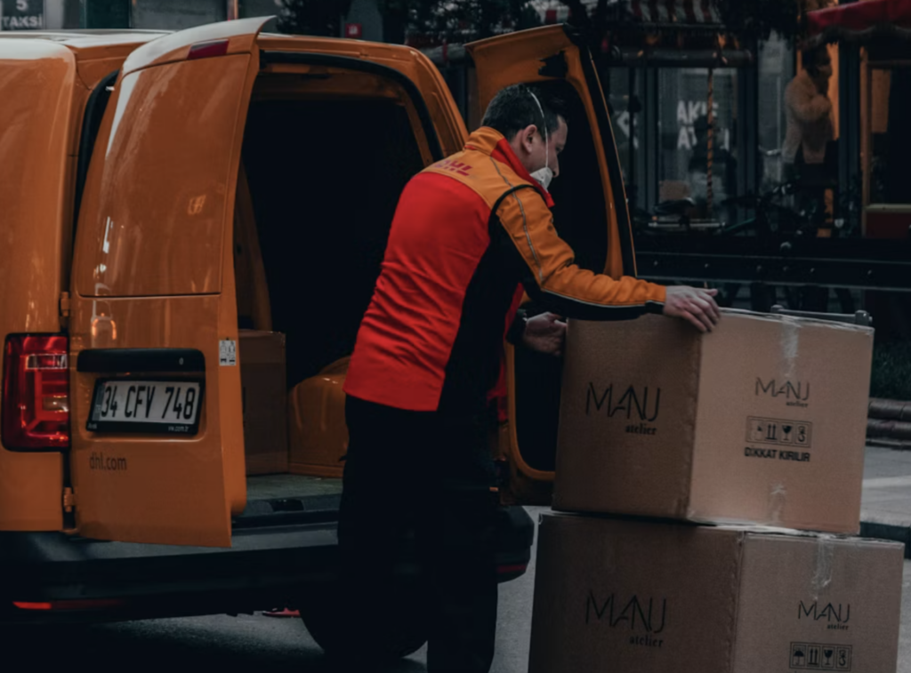 A delivery driver unloading cardboard boxes from the back of a van onto a street outside a store.