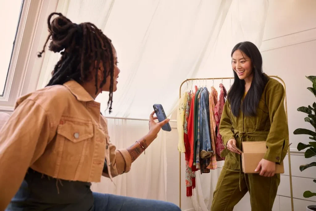 A woman films a friend on a phone beside a rail of clothing, holding a parcel.