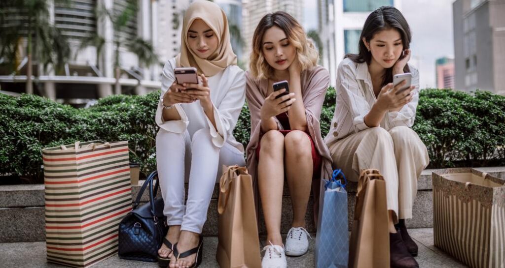 Three young shoppers sitting outside with shopping bags, each looking something up on their phones.