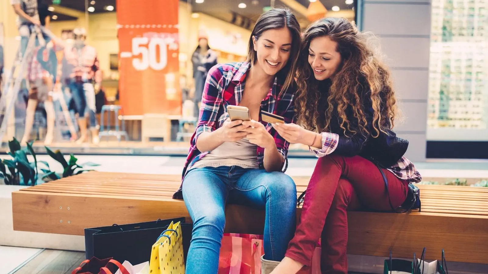 Two young women sitting in a shopping mall, checking a phone and holding a credit card and shopping bags.