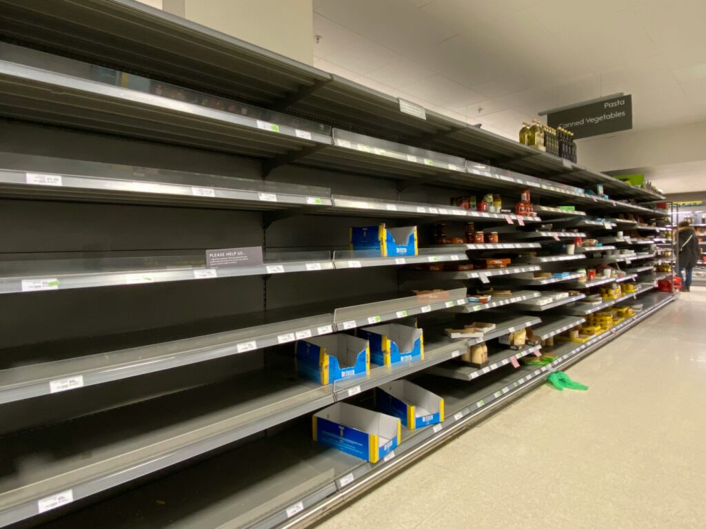 Empty supermarket shelves in a pasta and canned vegetables aisle, with a sign asking customers to limit purchases.