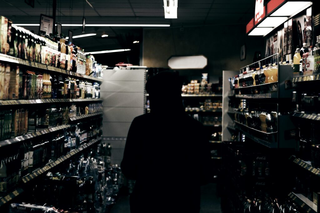 A shopper in silhouette standing in a dimly lit convenience store aisle between drinks shelves.