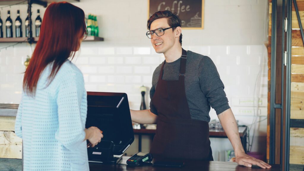 A smiling store associate in an apron talking with a customer at a counter beside a card reader and till.