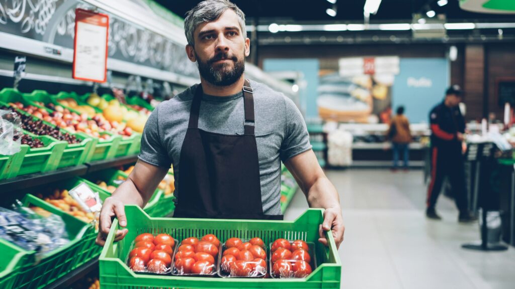 Grocery colleague carrying a crate of packaged tomatoes through the produce department.