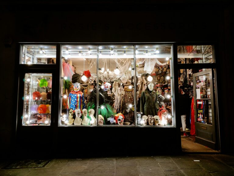 Lit store window at night filled with Halloween masks, costumes, wigs and cobweb decorations