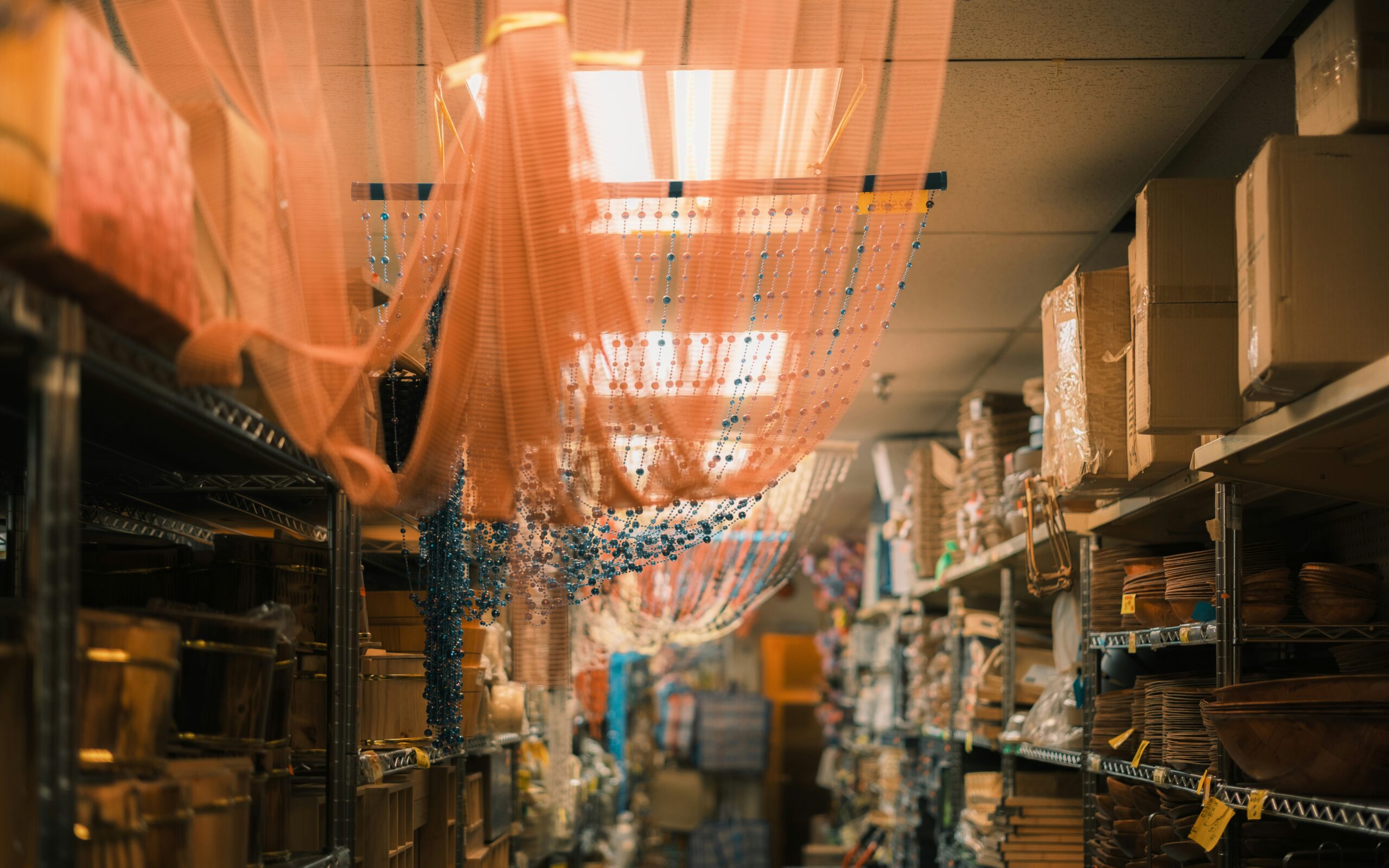 Packed store aisle with stocked shelves, cardboard boxes stacked overhead and hanging bead garlands
